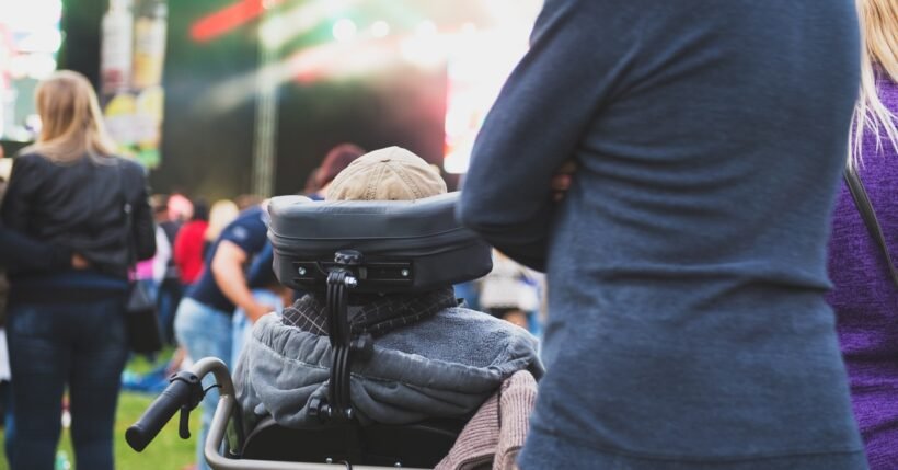 A person using a wheelchair watching a brightly lit outdoor festival stage while standing attendees gather nearby.
