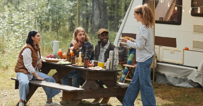 Three people are sitting at a wooden picnic table in the woods as a woman holds a plate of food. An RV is in the background.