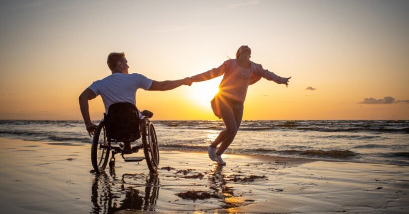 Two people moving along a beach with their hands together. One of them is running while the other is in a wheelchair.