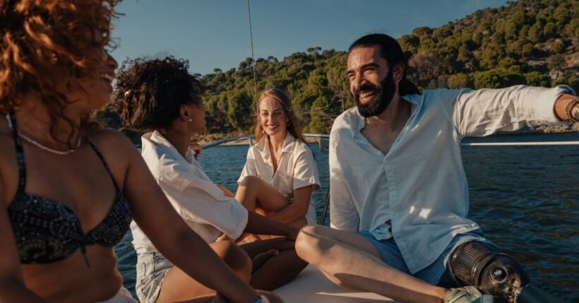 Four friends sit together on a boat, smiling and chatting, with blue water and a tree-covered shoreline behind them.